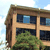 Pixelated image of a two-story building with brown trim, light tan walls, and dark blue windows, partially obscured by green foliage under a light blue sky.