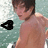 A shirtless person with short dark hair stands in front of a blurred water background, with a black triangular symbol on the left side of the frame.