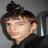 Pixelated static image of a person with dark hair styled in a high bun, wearing a dark top against a gray background.