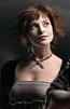 Static image of a woman with reddish-brown hair wearing a low-cut top and choker necklace against dark background.