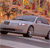 A silver sedan with visible headlights and wheels is positioned on a reflective surface under a colorful, blurred background of warm tones.