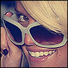 A close-up of a person with light brown hair wearing large white-framed sunglasses, holding the frame with their left hand while smiling broadly.