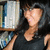 A person with long dark hair and a black sleeveless top stands in front of bookshelves, smiling slightly while the right side of their face is obscured by a gray rectangle.