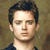 Pixelated portrait of a person with short dark hair, light skin, and a neutral expression, wearing a dark collared shirt against a muted brown background.