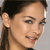 Pixelated close-up of a person's face with dark hair tied back, light skin, and a slight smile against a gray background.