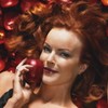A woman with voluminous red hair holds a shiny red apple near her face against a background of blurred red and brown shapes.
