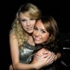 Two women embrace in a close-up pose against a dark background, one wearing a sleeveless silver dress with a textured pattern.