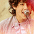 A person with curly brown hair wearing a light pink zip-up jacket sings into a silver microphone against a red and beige background.
