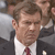 Pixelated close-up of a person in a dark suit, white shirt, and red tie, with brown hair and a neutral expression against a blurred background.