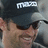 A pixelated image shows a person wearing dark sunglasses and a black cap with white text, smiling against a blurred background.