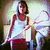 Pixelated image of a person in a white sleeveless top and red skirt seated with legs crossed, holding an object near their face.