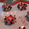 Aerial view of people gathered around a red circular table with a white symbol, surrounded by red and gray architectural elements, with 'EAST HIGH' text and heart symbols.