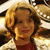 Pixelated static image of a person with curly hair wearing a red top and light-colored jacket against a blurred background.