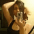 A person with long brown hair wearing a black tank top holds a silver digital camera up to their face, arms raised behind head, in a four