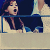 A pixelated image shows a person with long brown hair wearing a white top, mouth open in a shout or yell, with another person in a white patterned shirt partially visible behind them.