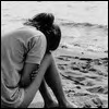 Black-and-white image of two people embracing on a beach, one with dark hair tied up, both wearing light-colored shirts, facing the water.