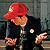 Pixelated figure wearing red cap, black jacket with white pocket square, and dark pants; hands raised in gesture against blurred background.