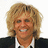 Pixelated portrait of a person with light brown curly hair, wearing dark jacket and white shirt, smiling broadly against plain background.