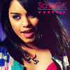 A pixelated close-up of a person with long dark hair wearing a colorful patterned top and a red garment underneath, with visible neck tattoo and pink text overlay in the upper right corner.