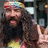 Pixelated static image of a person with long dark hair and beard wearing colorful headband, black top, and yellow garment against blurred background.