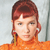 Pixelated image of a person with reddish-brown hair styled in a high bun, wearing an orange turtleneck sweater against a light gray background.
