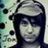 Pixelated static image of a person with dark hair and light skin wearing a cap, set against a patterned background.