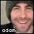 Pixelated close-up of a smiling person with light skin, dark hair, and visible teeth; black clothing on the left shoulder; white text 'adam' at bottom-left.