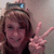 A person with light brown hair makes a peace sign, wearing a dark top and headband with blue text.