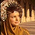 Pixelated static image of a person with curly hair and warm-toned clothing against a dark background, appearing to be from an older digital media source.