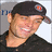 Pixelated image of a person wearing dark clothing and a black cap with red-and-gray emblem, smiling slightly against light background.