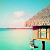 Pixelated beach house with thatched roof over turquoise water under light blue sky with scattered clouds.