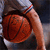 A hand holds a red basketball with visible seams against a blurred background of dark and light tones.