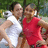 Two people stand close together outdoors, one in a white sleeveless top and the other in a red sleeveless dress, with green foliage background.