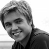 Black-and-white photo of a person with short light hair wearing a dark collared shirt, smiling with visible teeth against a bright background.