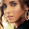 A close-up of a person with light brown wavy hair, blue eyes, and a gold hoop earring, wearing a dark top with a light-colored collar.