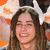 A woman with long brown hair and a white headband smiles broadly, wearing an orange top against a blurred background.