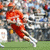 Pixelated image of a person in red clothing holding a lacrosse stick on grass with blurred background, next to a black-and-white skull and crossbones symbol.