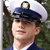 A person wearing a dark blue peaked cap with a light-colored brim and a circular emblem on top, standing against a blurred green background.