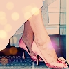 A close-up of a person's lower legs and feet wearing pink high-heeled shoes with a small flower detail, standing on a blue checkered surface.