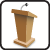 Pixelated brown podium with a microphone on top, set against a white background within a black border.