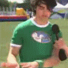 A person in a green shirt with a logo holds a black microphone, standing outdoors with a blurred background.