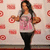 A woman in a pink graphic t-shirt and black pants stands against a red-and-white patterned backdrop with circular logos, holding two brown objects.