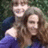 Two figures in close embrace, one with light hair and blue clothing, the other with brown hair and dark clothing against a blurred green background.