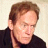Static pixelated image of a person's head and shoulders in dark clothing against light background, with visible dithering and low-resolution rendering artifacts.