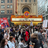 A pixelated image shows a crowd gathered in front of a building with a large marquee sign displaying a blank white rectangle, surrounded by a golden-brown border.