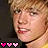 Pixelated close-up of a person with light brown hair and fair skin, featuring three pink heart icons in the bottom-left corner on a dark background.