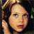 Pixelated close-up of a person with light brown hair and fair skin, wearing dark clothing against a dark background.