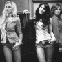 Three women pose in a ; left woman wears gray top and shorts, middle woman white shirt and jeans, right woman red top. Left woman's outfit changes from grayscale to color
