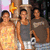 Three people stand together in a group photo; the person on the left wears a yellow dress, the middle person wears a gray tank top and white shorts, and the right person wears a brown shirt with light blue jeans.