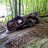 Pixelated static image shows dark vehicle on textured ground with green foliage background.
